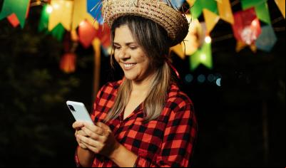 mulher branca com trajes de festa junina segurando e olhando para o celular sorrindo - curiosidades sobre o São João - Assaí Atacadista